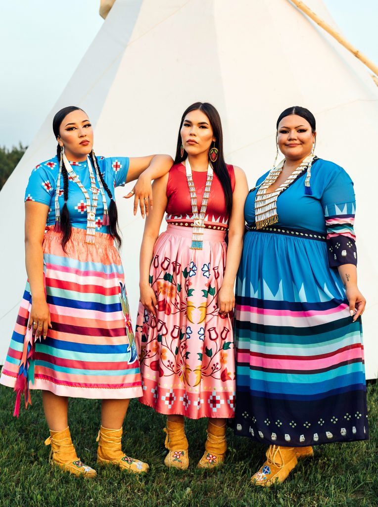 three woman in  Maskawitehew designs standing on green grass with a tipi behind them
