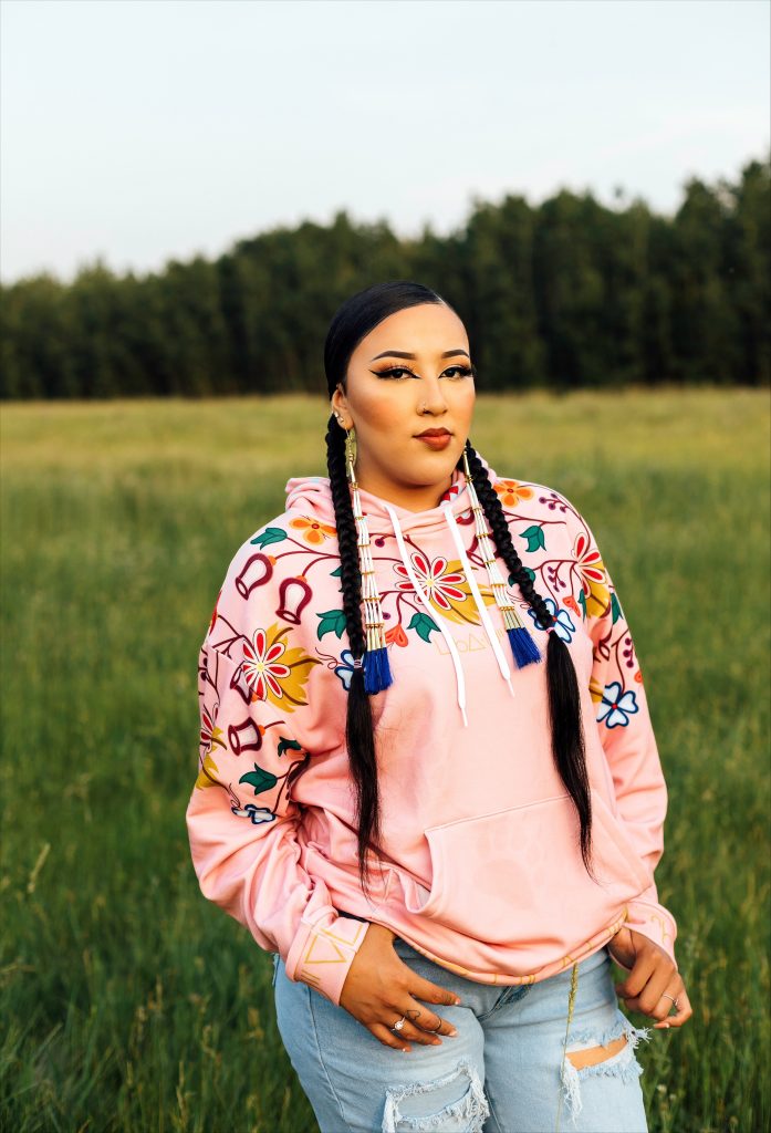 woman in Maskawitehew design cheyenne pink hoodie standing on green grass with trees behind her